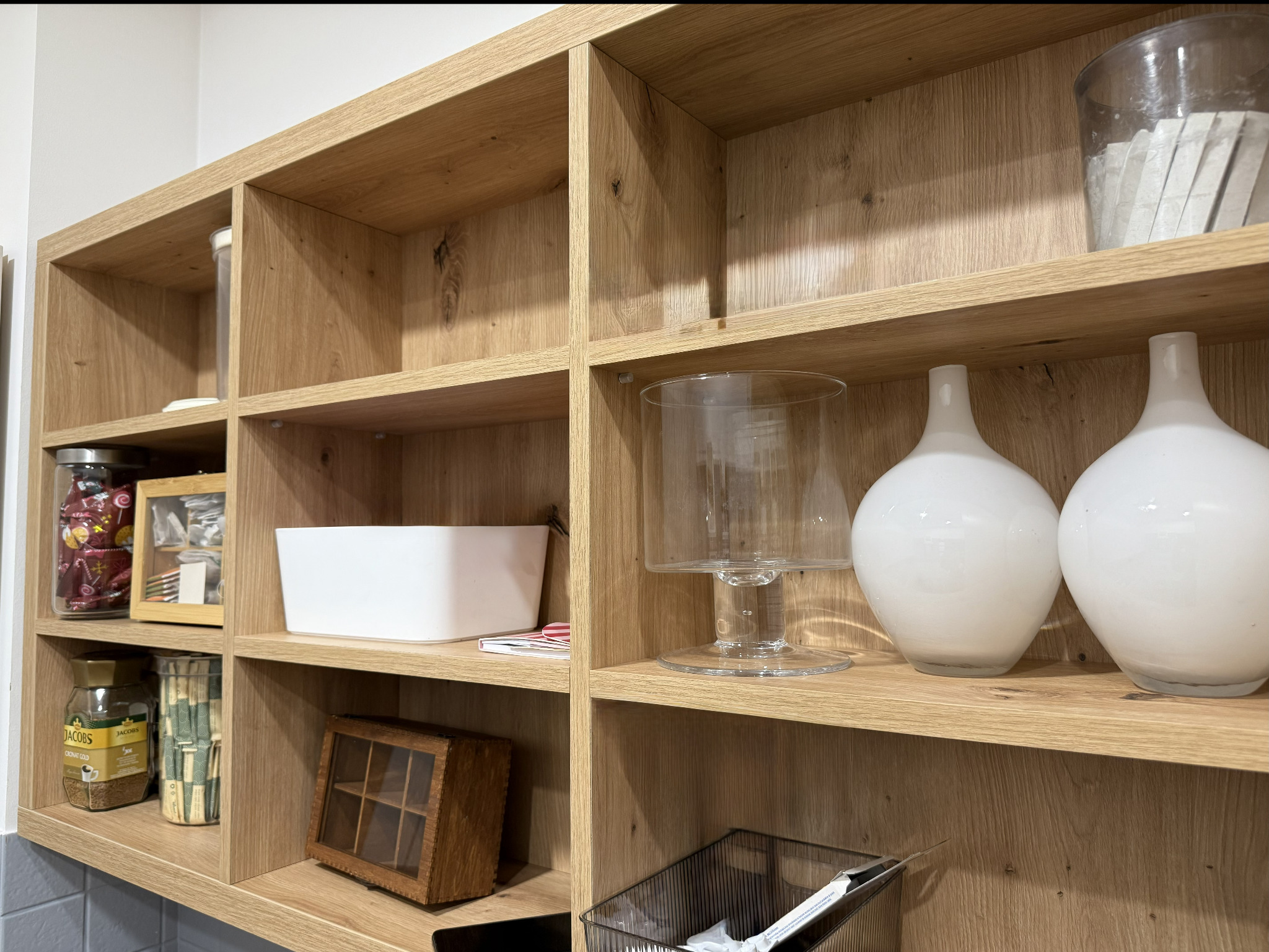 Półki z jasnego drewna z dekoracyjnymi przedmiotami i pojemnikami. Widoczne słoiki z kawą, pudełka na herbatę i białe wazony. Naturalne oświetlenie, ciepła kolorystyka.