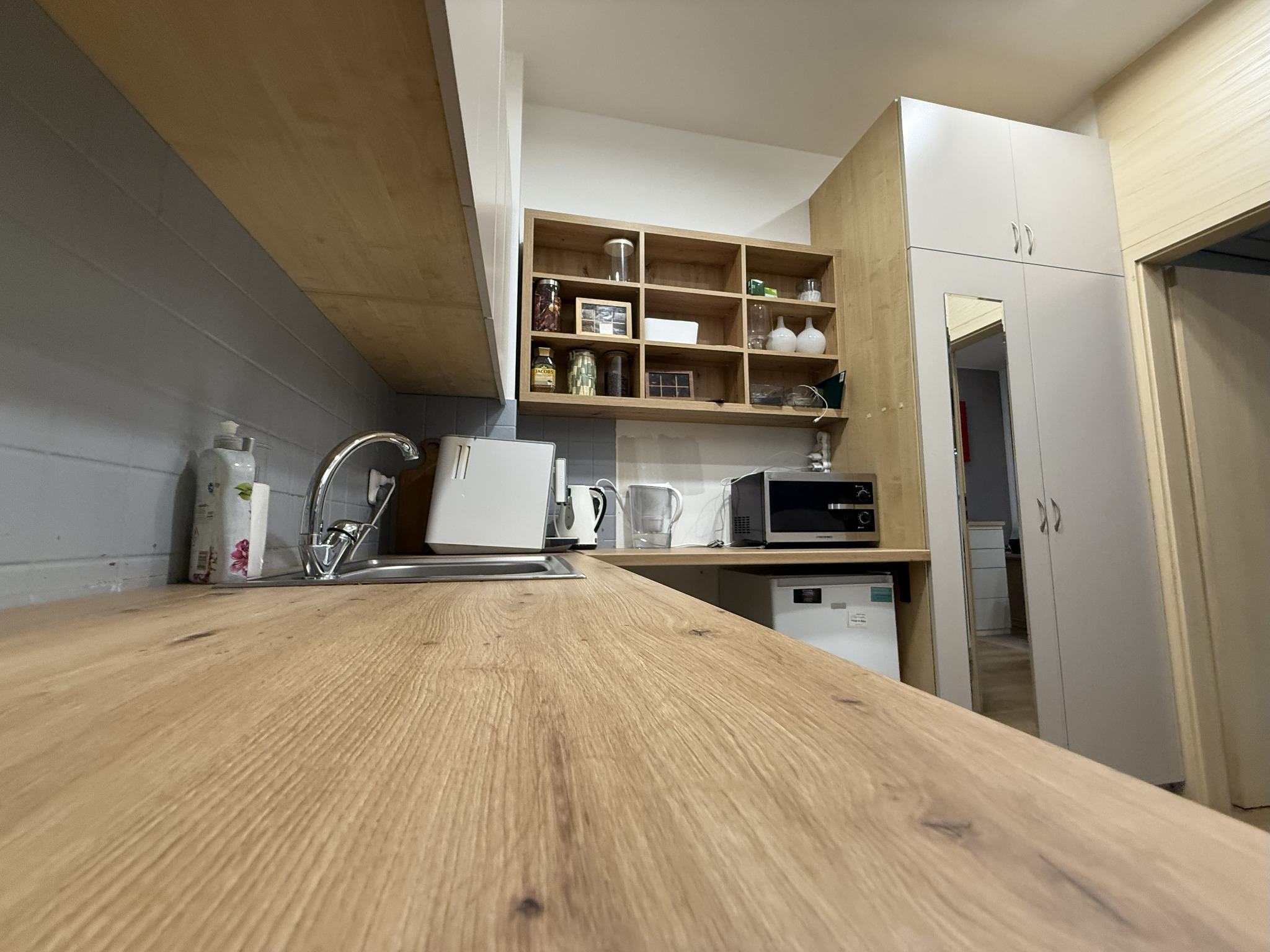 Jasna kuchnia z drewnianym blatem, stalowym zlewem i białymi szafkami. Na półkach dekoracje i sprzęty kuchenne. Widok z perspektywy blatu, podkreślający jego strukturę.