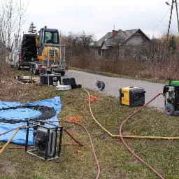 Prace ziemne z użyciem minikoparki i agregatów prądotwórczych przy drodze, na trawie rozłożona niebieska plandeka z czarnym kablem w zwojach, w tle budynek mieszkalny i drzewa bez liści.