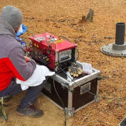 Pracownik w szarej bluzie z czerwonymi wstawkami, siedzący na składanym krześle, obsługuje czerwoną maszynę z manometrem i wyświetlaczem cyfrowym, umieszczoną na skrzyni z kółkami; obok leżą...