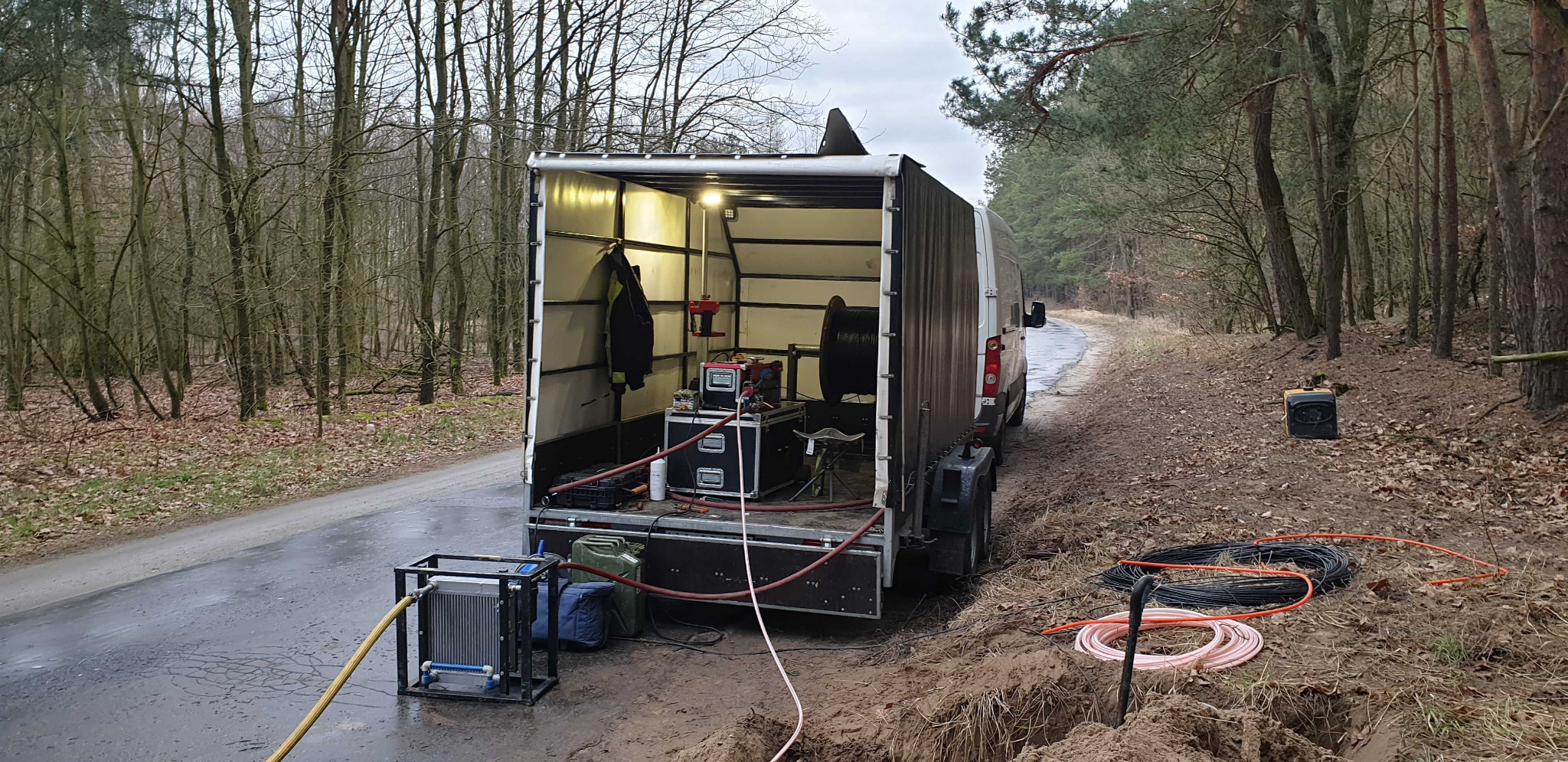 Mobilne stanowisko pracy w przyczepie na skraju lasu, sprzęt pomiarowy i zwój kabli światłowodowych na ziemi, przygotowanie do instalacji telekomunikacyjnej.