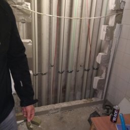 Instmar - Ściana z pionowymi rurami instalacyjnymi, częściowo osadzonymi w murze, z widocznymi przewodami i uchwytami mocującymi, w trakcie prac remontowych, z widocznym fragmentem sylwetki osoby