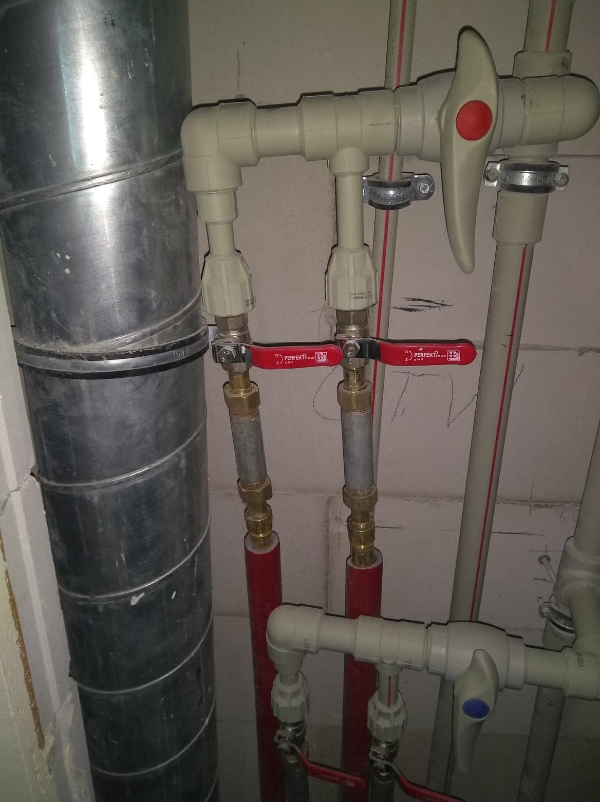 Fragment instalacji z rur z tworzywa sztucznego, zaworów z czerwoną rączką i metalowej rury wentylacyjnej na tle surowej ściany z betonu komórkowego.