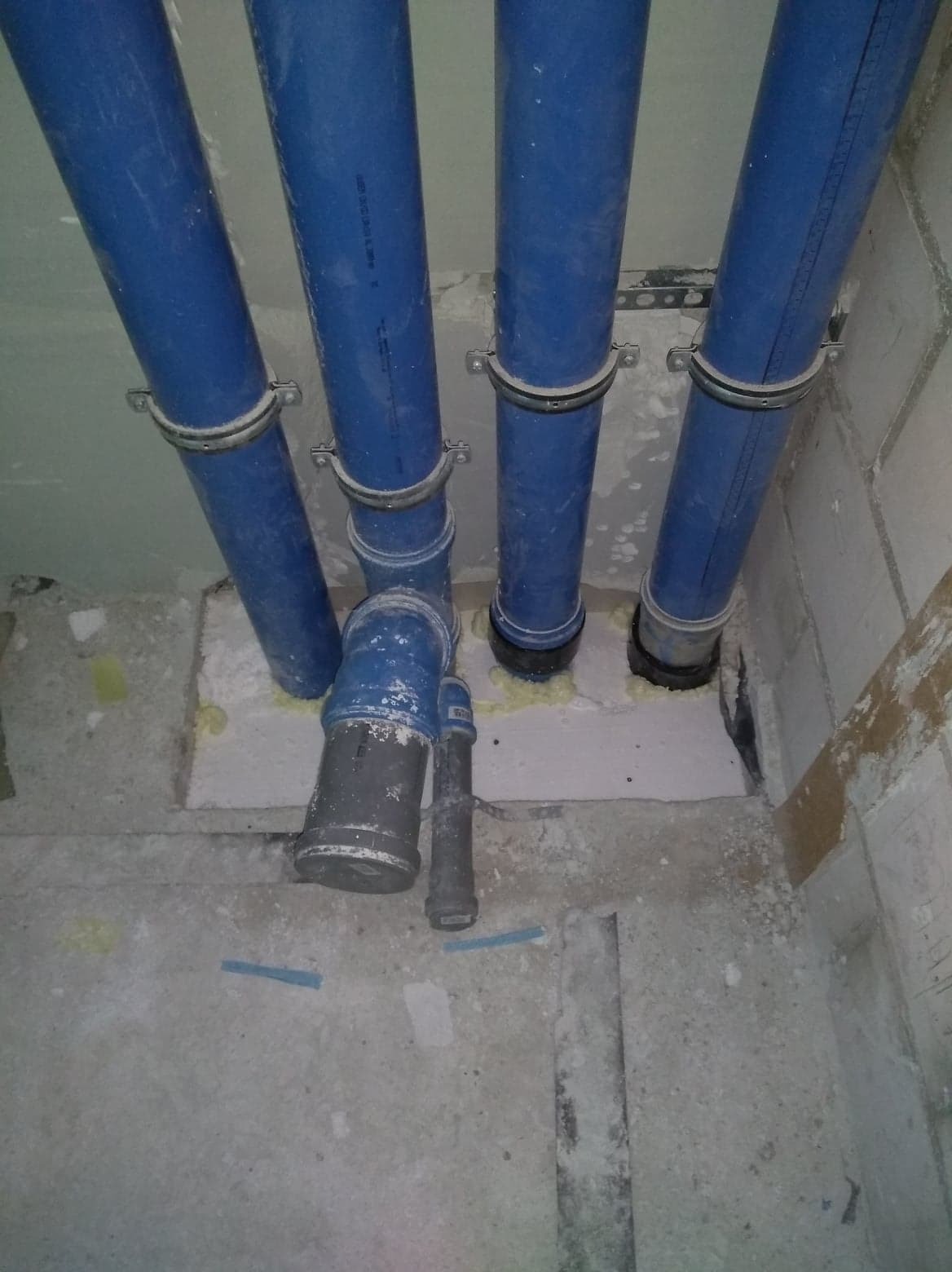 Pionowe, niebieskie rury kanalizacyjne z metalowymi obejmami, w surowym stanie deweloperskim, widoczne fragmenty pianki montażowej i przewody.