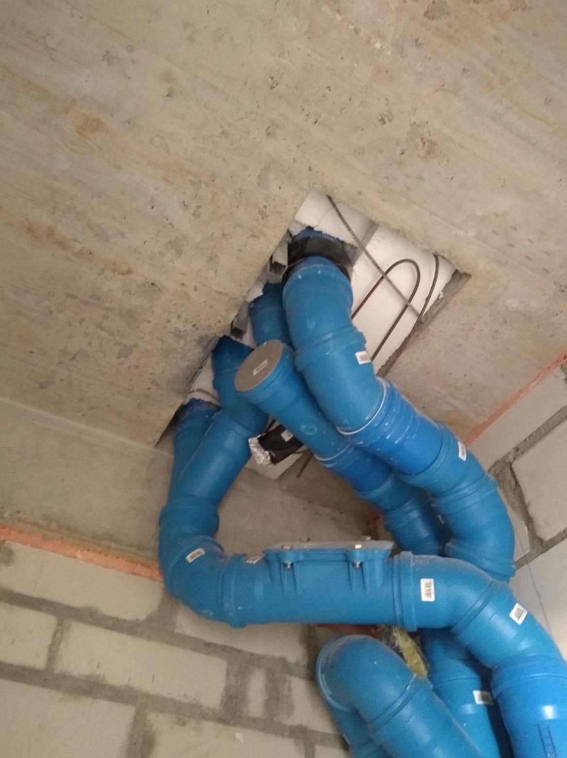 Niebieskie rury kanalizacyjne z tworzywa sztucznego prowadzone przez otwór w surowym betonowym stropie, widoczne elementy izolacji termicznej.