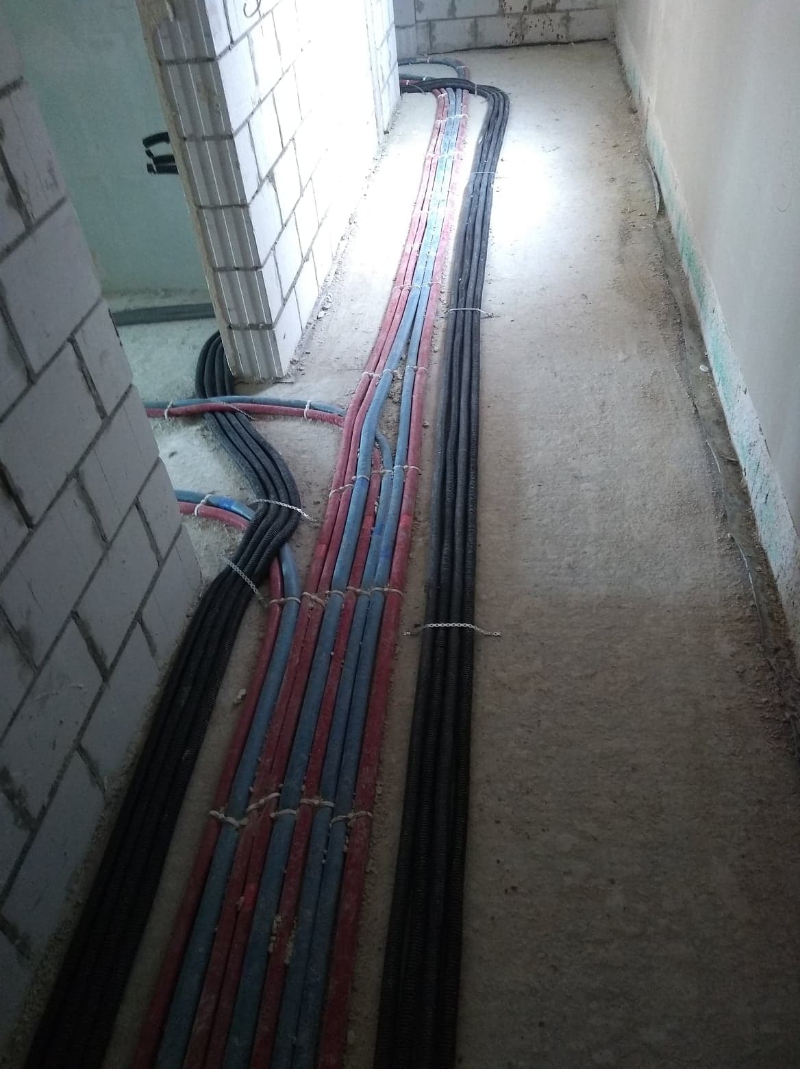 Wiązki rur instalacyjnych w kolorach czerwonym i niebieskim oraz czarne rury karbowane ułożone równolegle na surowym betonowym podłożu w trakcie prac instalacyjnych w budynku w stanie deweloperskim.