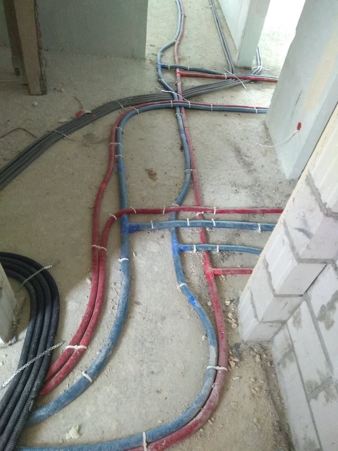 Wiązki rur instalacyjnych w kolorze niebieskim, czerwonym i szarym, prowadzone po surowej podłodze w trakcie budowy, zabezpieczone białymi opaskami zaciskowymi, widoczne fragmenty ścian z betonu...