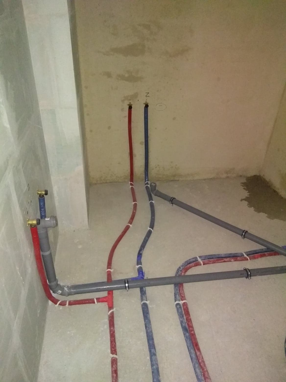 Surowe wnętrze łazienki w trakcie prac hydraulicznych: widoczne rury z tworzywa w kolorach czerwonym, niebieskim i szarym, prowadzone po podłodze i ścianach, z licznymi złączkami i obejmami...