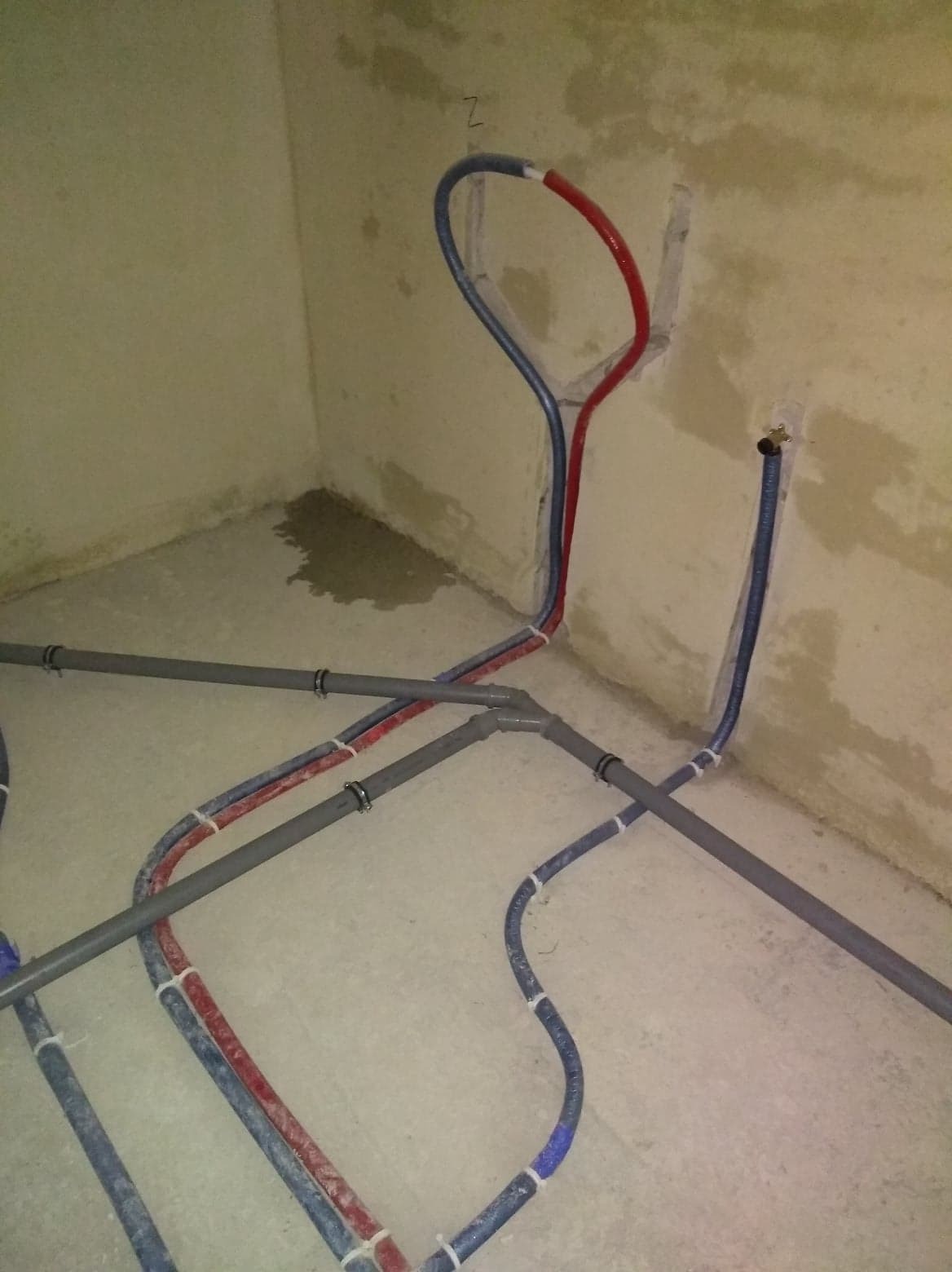 Wewnętrzne rury wodne i kanalizacyjne, częściowo przymocowane do ściany, z widocznymi złączkami i opaskami zaciskowymi, w surowym stanie wykończenia pomieszczenia.