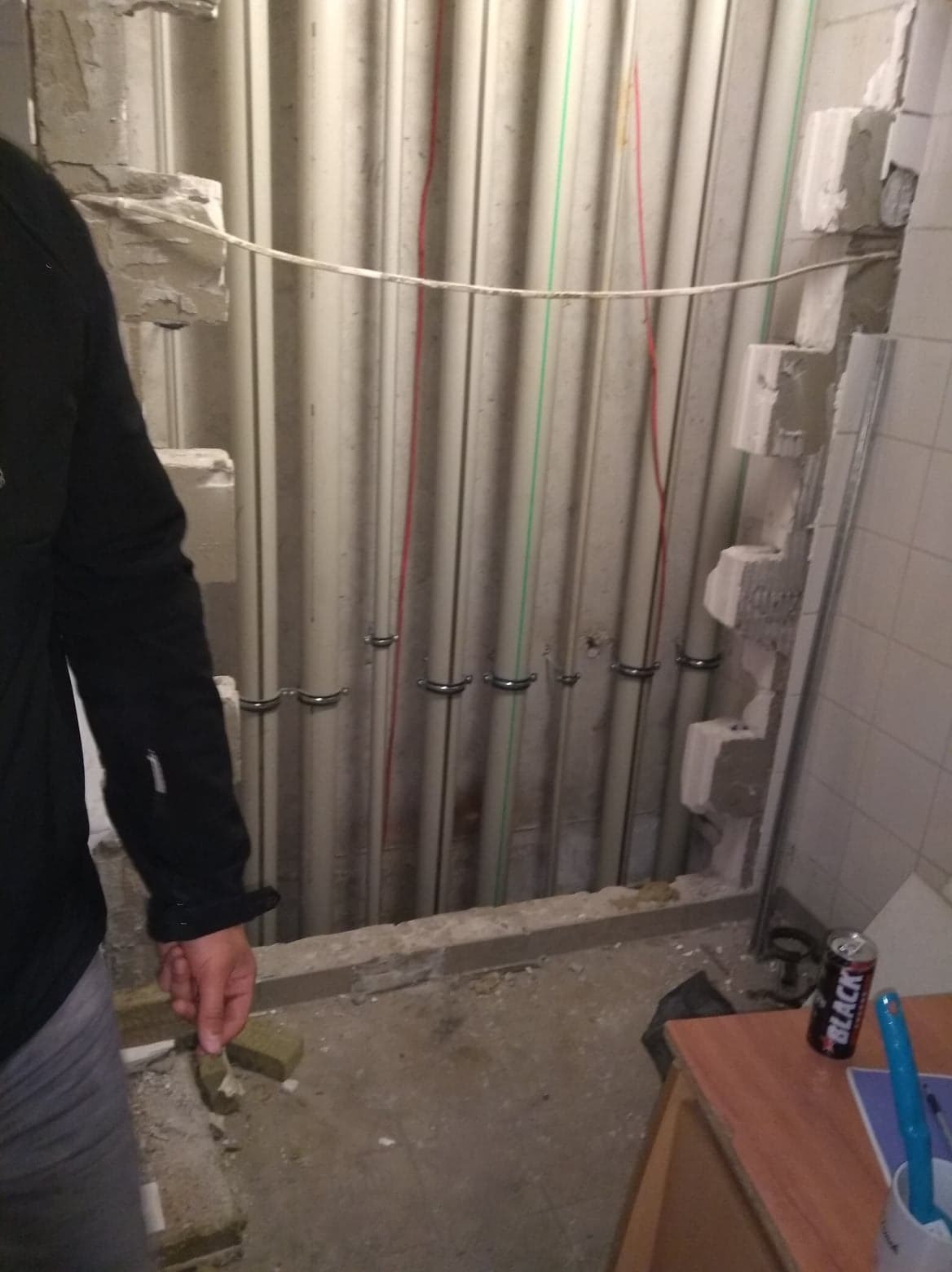 Ściana z pionowymi rurami instalacyjnymi, częściowo osadzonymi w murze, z widocznymi przewodami i uchwytami mocującymi, w trakcie prac remontowych, z widocznym fragmentem sylwetki osoby