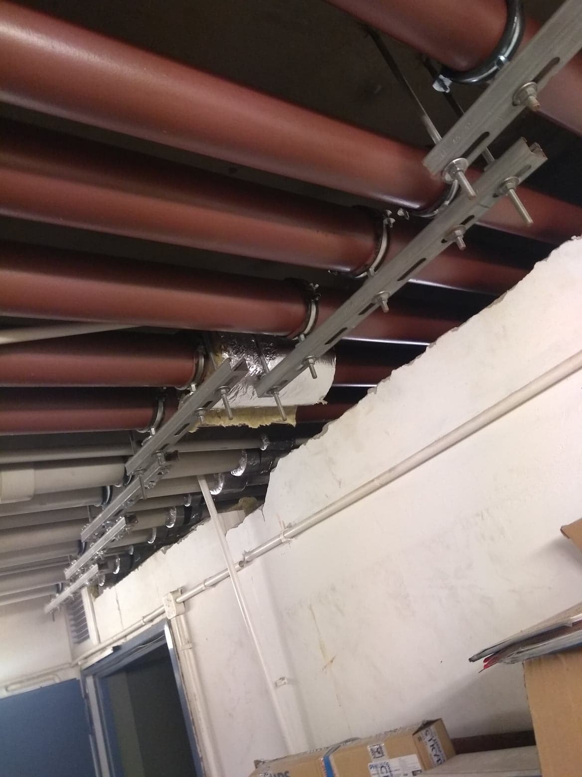 Brązowe rury pod sufitem, mocowane za pomocą metalowych uchwytów, widoczne elementy izolacji i uszkodzony sufit.