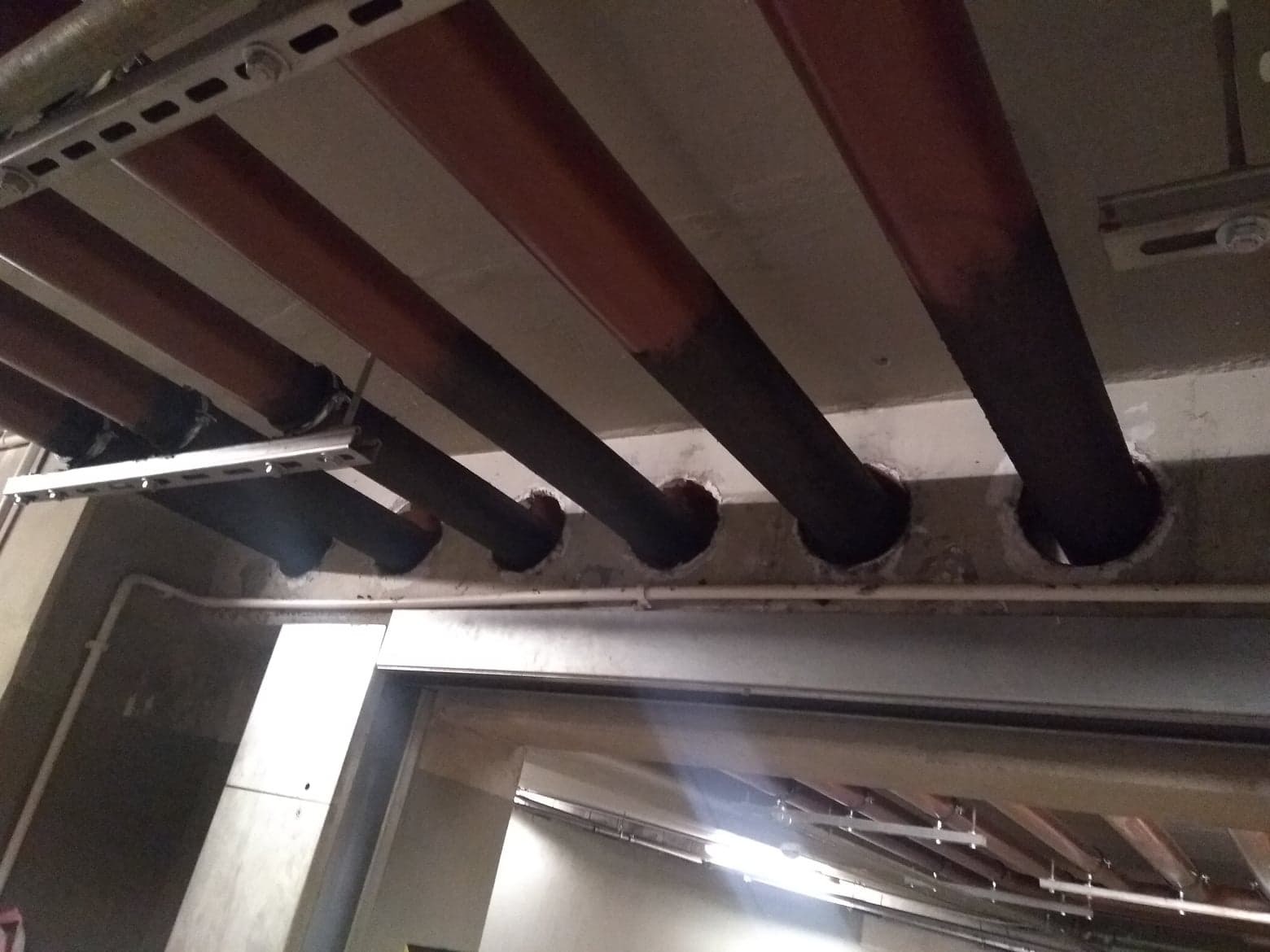 Widok z dołu na rzędy rur przechodzących przez otwory w suficie, niektóre z nich mają ślady rdzy, a otwory są niedokładnie wykończone.