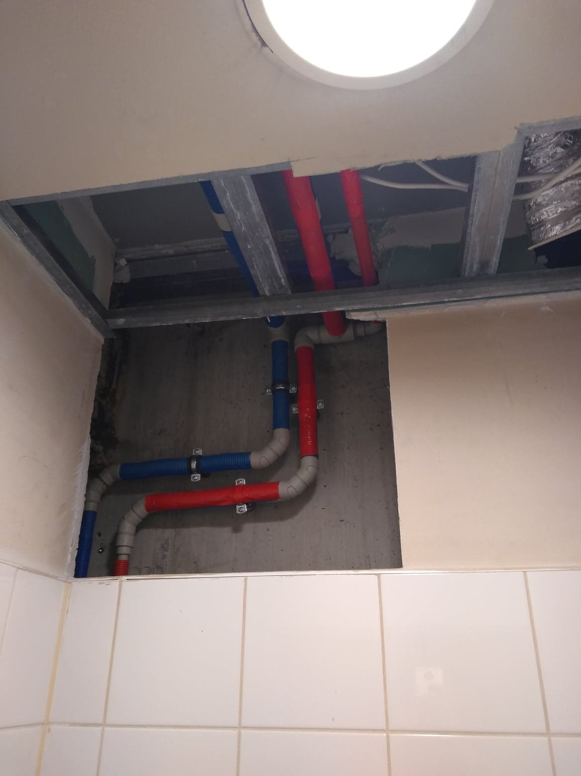 Widok na sufit podwieszany w łazience w trakcie prac z instalacją wodną, widoczne rury w kolorach niebieskim i czerwonym, mocowane do ściany za pomocą metalowych uchwytów, białe płytki na dolnej...