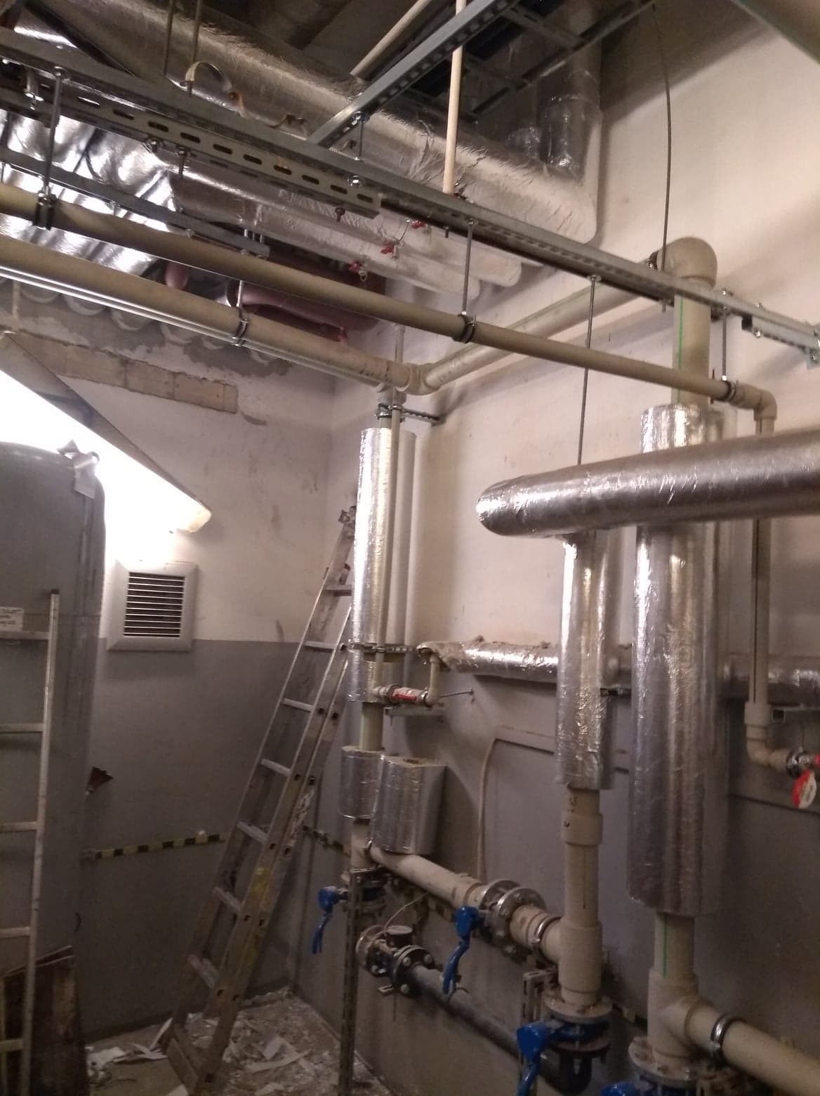 Widok na instalację rur z izolacją termiczną, zaworami i drabinką w pomieszczeniu technicznym o surowym wykończeniu.
