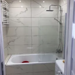 Wykończona łazienka z wanną obudowaną płytkami imitującymi marmur, szklanym parawanem nawannowym, grzejnikiem drabinkowym i pralką.