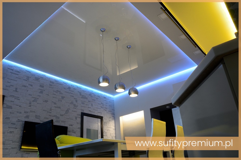 Biały, podwieszany sufit z oświetleniem LED w kolorze niebieskim i trzema srebrnymi lampami wiszącymi, widok z dołu na jadalnię z elementami kuchni w tle.