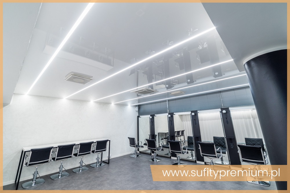 Nowoczesne wnętrze salonu fryzjerskiego z białym, błyszczącym sufitem podwieszanym z liniowym oświetleniem LED, czarnymi fotelami fryzjerskimi i stanowiskami z lustrami.