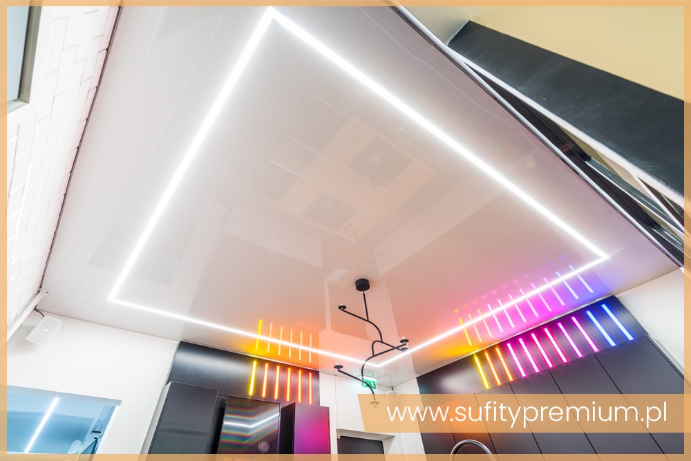 Biały, błyszczący sufit napinany z kwadratowym obramowaniem z białego światła LED, dodatkowo podświetlenie RGB nad czarnymi szafkami kuchennymi i nowoczesna lampa wisząca.
