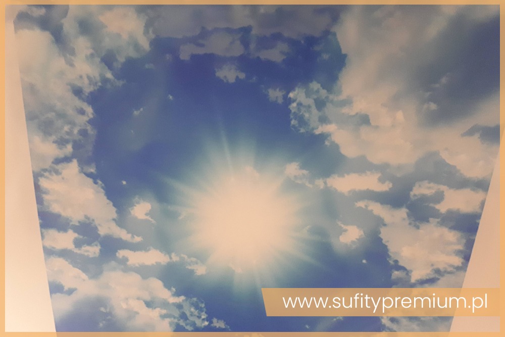 Niebieskie niebo z chmurami i centralnie umieszczonym słońcem, imitacja sufitu napinanego z adresem strony internetowej w dolnej części kadru.