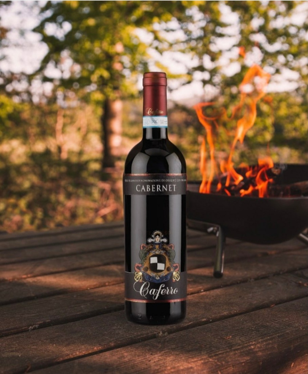Butelka czerwonego wina Cabernet Caferro na drewnianym stole piknikowym, z rozpalonym grillem w tle i zielenią drzew