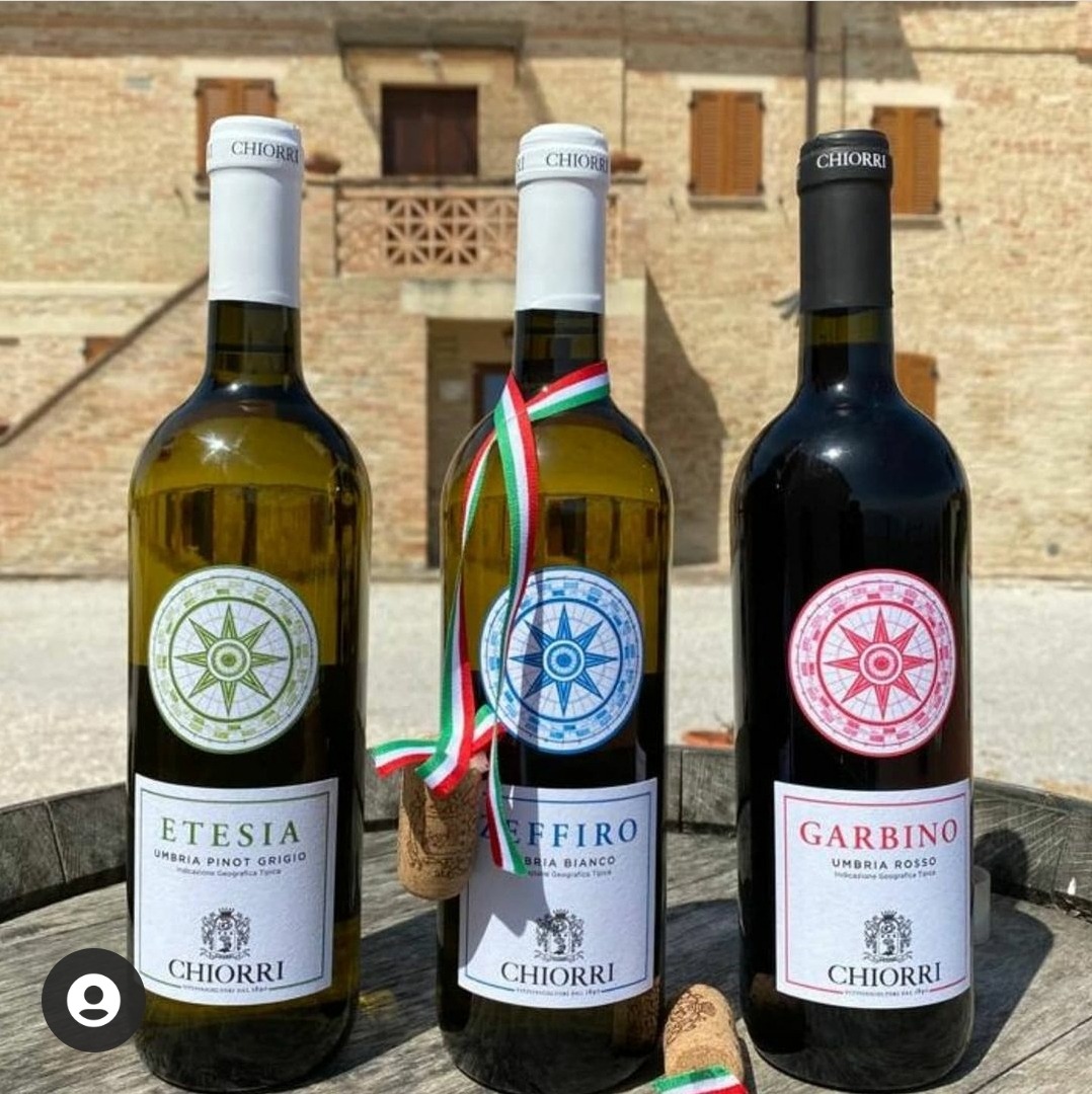 Trzy butelki wina Chiotti: białe, z zieloną etykietą, białe z wstążką w kolorach włoskiej flagi i korek, oraz czerwone, na tle ceglanego budynku.