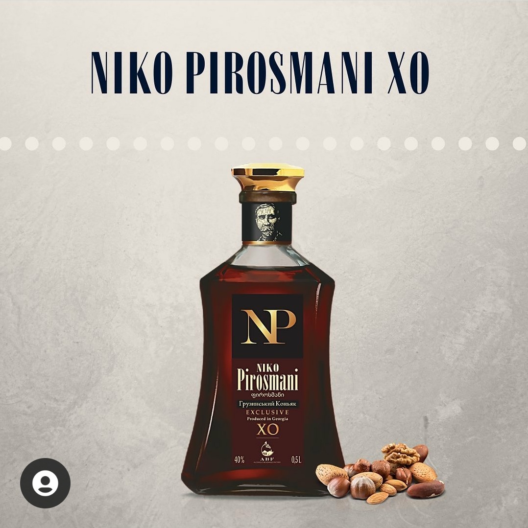 Butelka gruzińskiego koniaku Niko Pirosmani XO o nietypowym kształcie, z portretem na szyjce, stoi obok orzechów włoskich i laskowych na neutralnym tle.