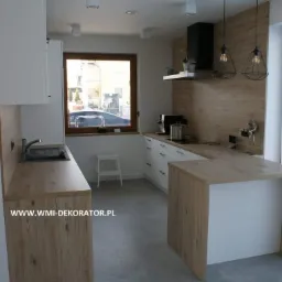 Nowoczesna kuchnia z drewnianymi blatami i białymi szafkami, oświetlona wiszącymi lampami w stylu industrialnym, widok na okno z zewnątrz.