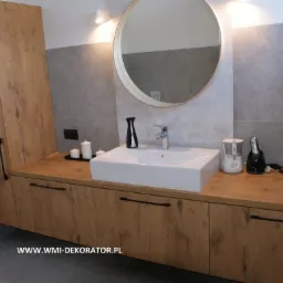 Nowoczesna łazienka z drewnianą szafką pod umywalką, okrągłym lustrem i szarą płytką na ścianie, oświetlona ciepłym światłem.