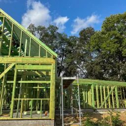 Konstrukcja domu w systemie szkieletowym, impregnowane drewno w odcieniach zieleni, widoczny fragment rusztowania i pracownik na wysokości, błękitne niebo w tle.