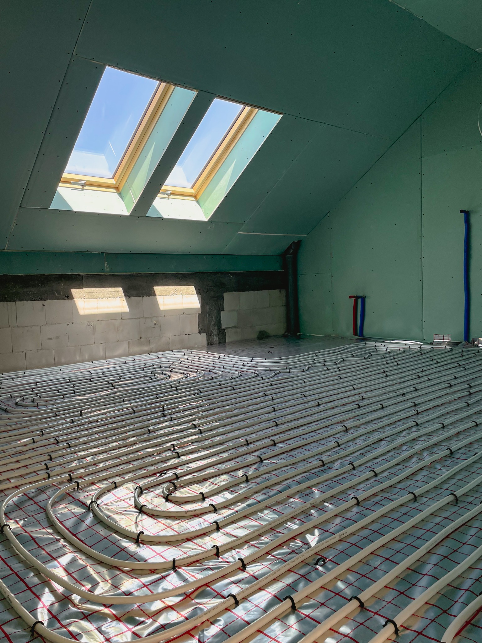 Instalacja ogrzewania podłogowego w surowym wnętrzu poddasza z oknami dachowymi i ścianami z płyt gipsowo-kartonowych.