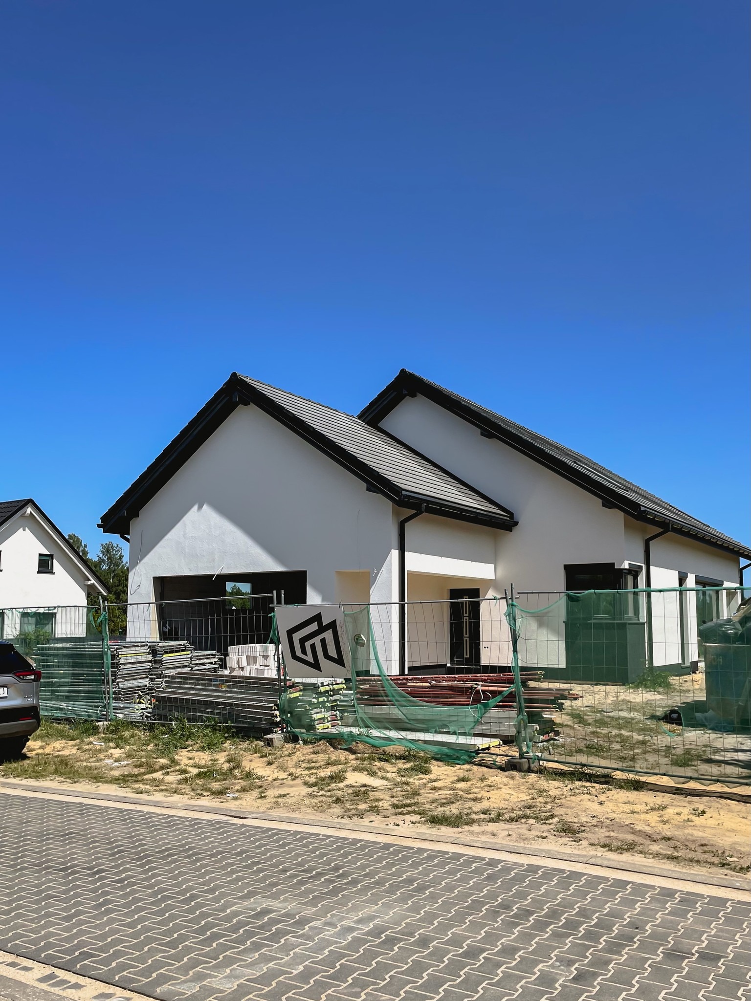 Biały, parterowy budynek w trakcie budowy, z czarnym dachem i stolarką okienną; widoczne rusztowania i materiały budowlane na posesji, błękitne niebo w tle.