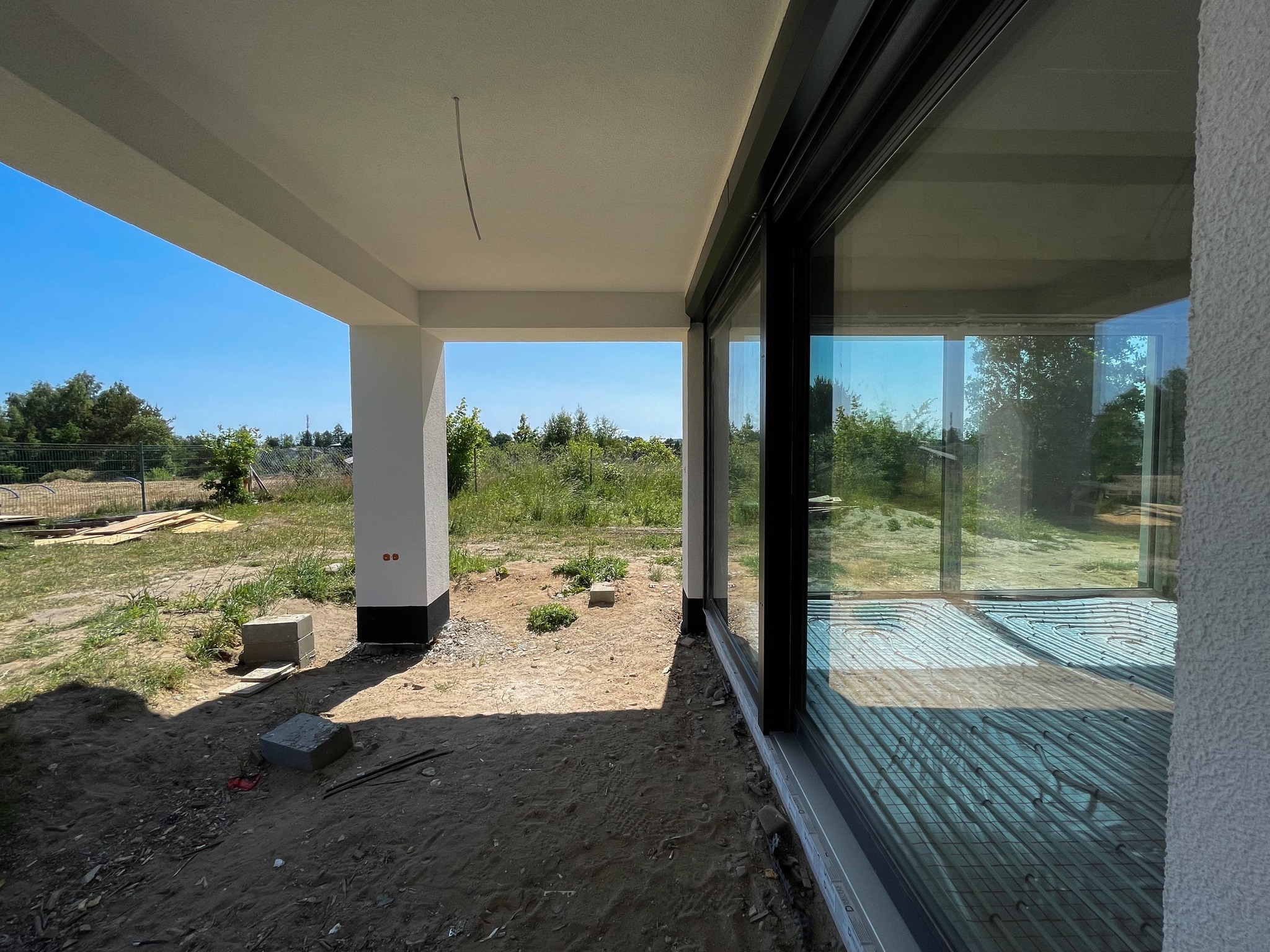 Wnętrze domu w trakcie budowy, widoczne ogrzewanie podłogowe i duże okno z widokiem na zewnątrz, surowy stan wykończenia.