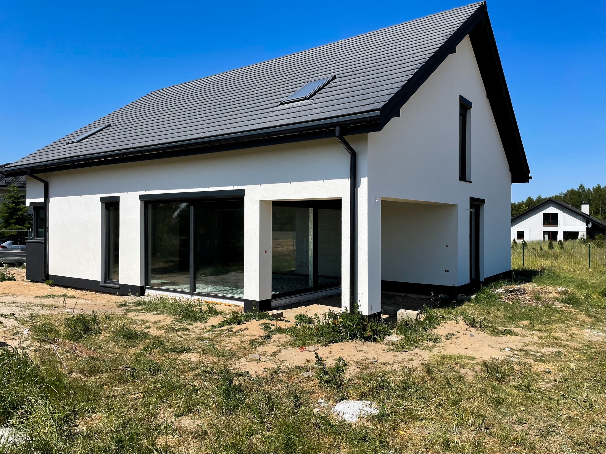 Nowoczesny, parterowy budynek z białymi ścianami i grafitowym dachem, duże przesuwne okna, widok na budowę w stanie surowym otwartym, na trawniku z piaskiem i chwastami.