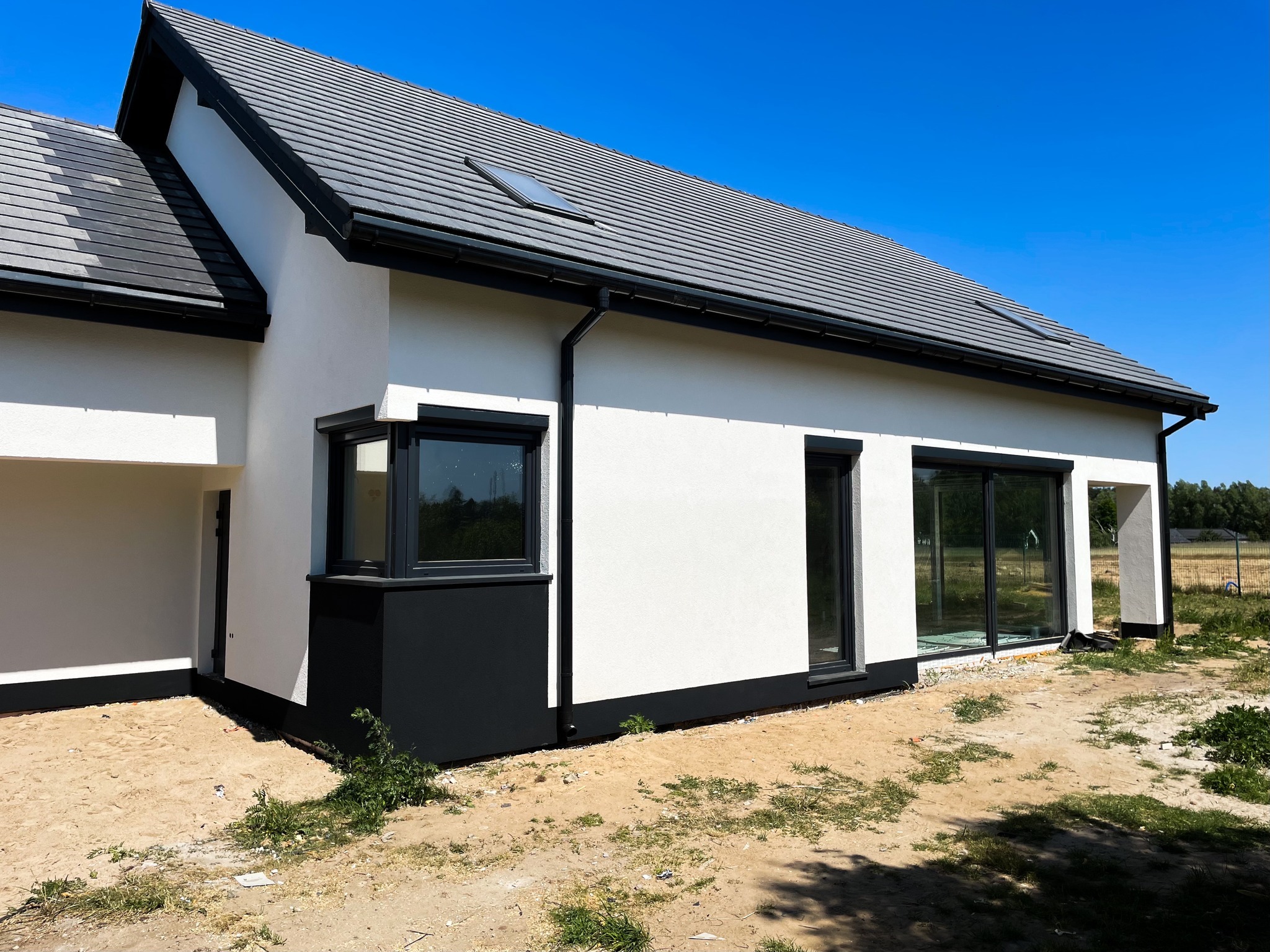 Nowoczesny, parterowy budynek z białymi ścianami i grafitową dachówką, z oknem dachowym, na etapie wykończenia terenu wokół budynku.