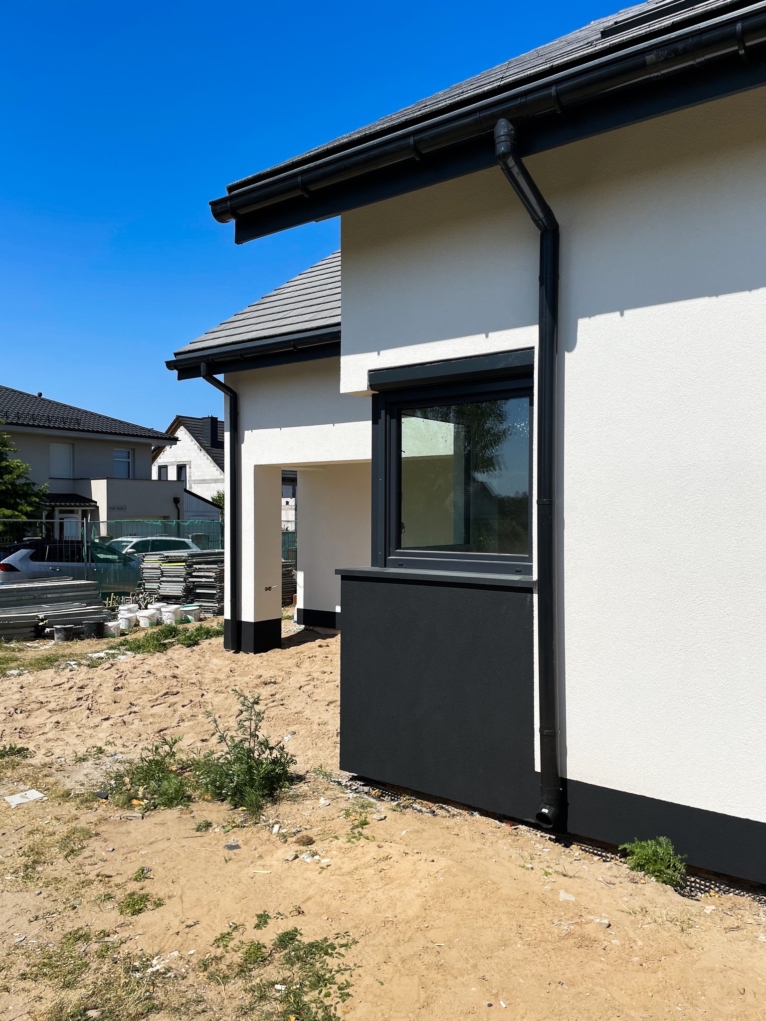 Fragment elewacji domu w trakcie budowy, widoczne orynnowanie, okno z czarną ramą i stos materiałów budowlanych w tle.