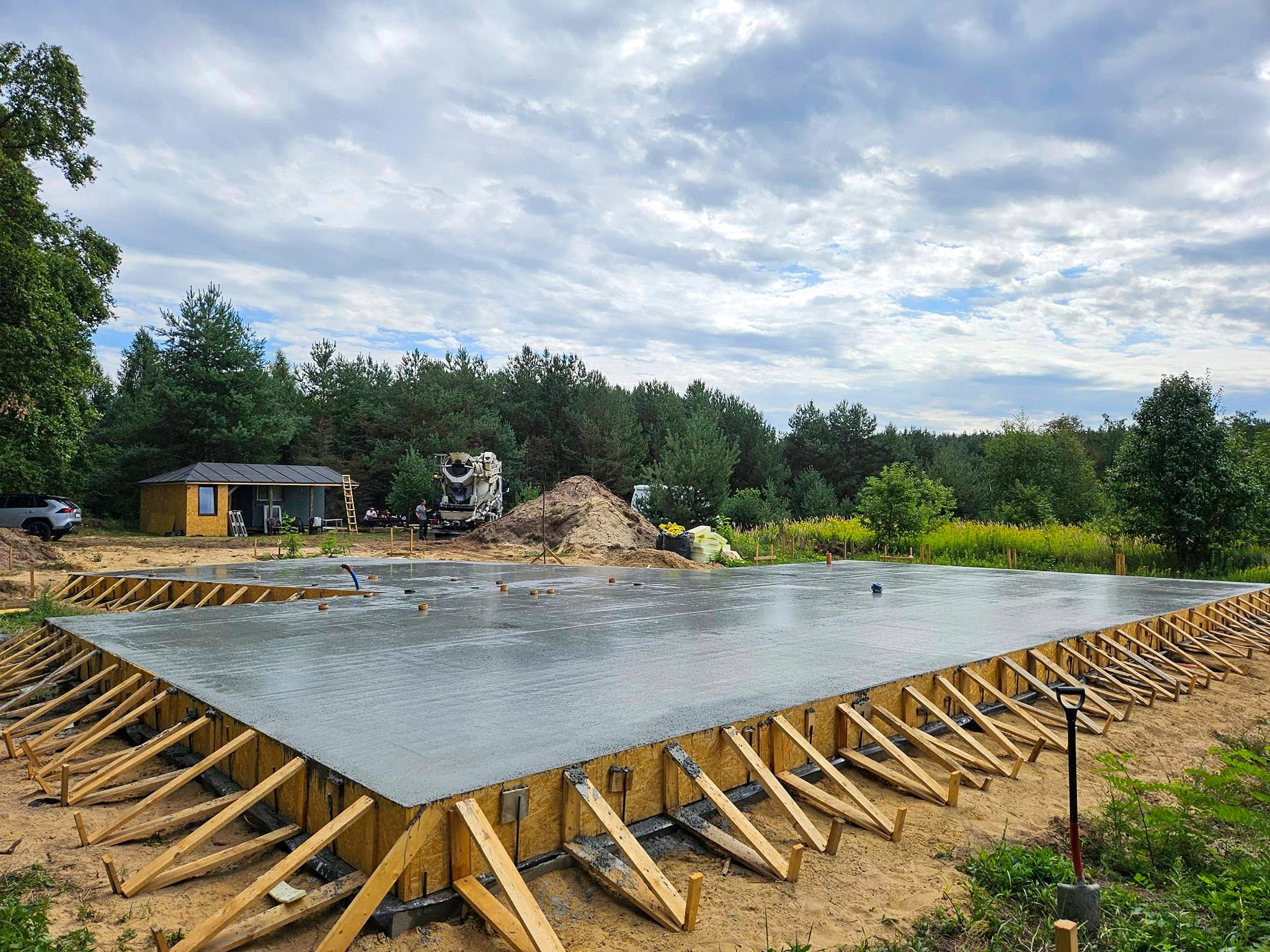 Wylany fundament pod budowę, z widocznym szalunkiem drewnianym i betoniarką w tle, otoczony zielenią.