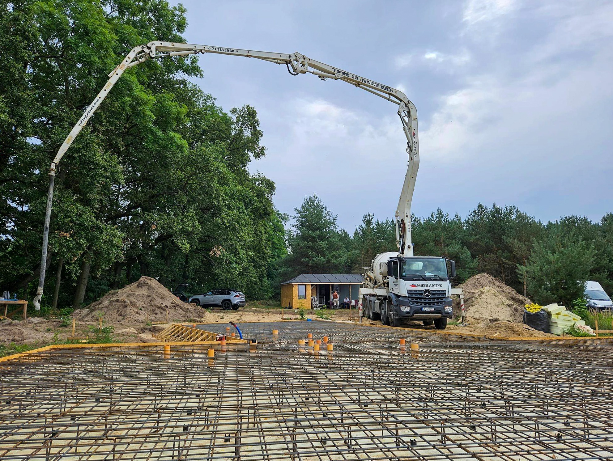 Zbrojenie fundamentów pod budowę domu na działce z pompą do betonu i małym domkiem w tle.