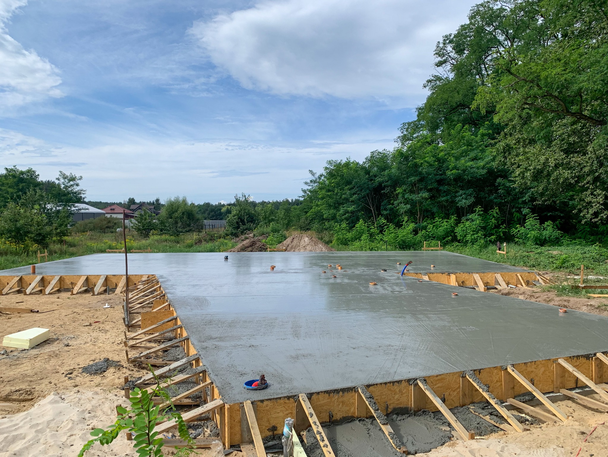 Wylany fundament pod budowę domu, szalunek z desek, widoczne instalacje podposadzkowe, tło: drzewa i błękitne niebo z chmurami.