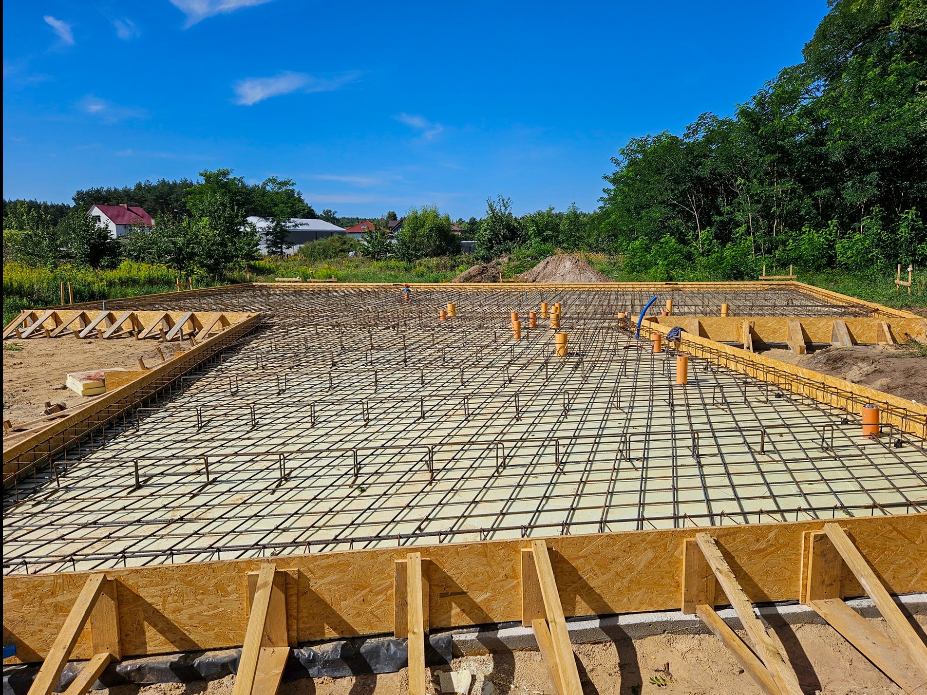 Przygotowany fundament z deskowaniem i zbrojeniem pod budowę, widoczne pomarańczowe rury instalacyjne, w tle zabudowa jednorodzinna i zieleń.