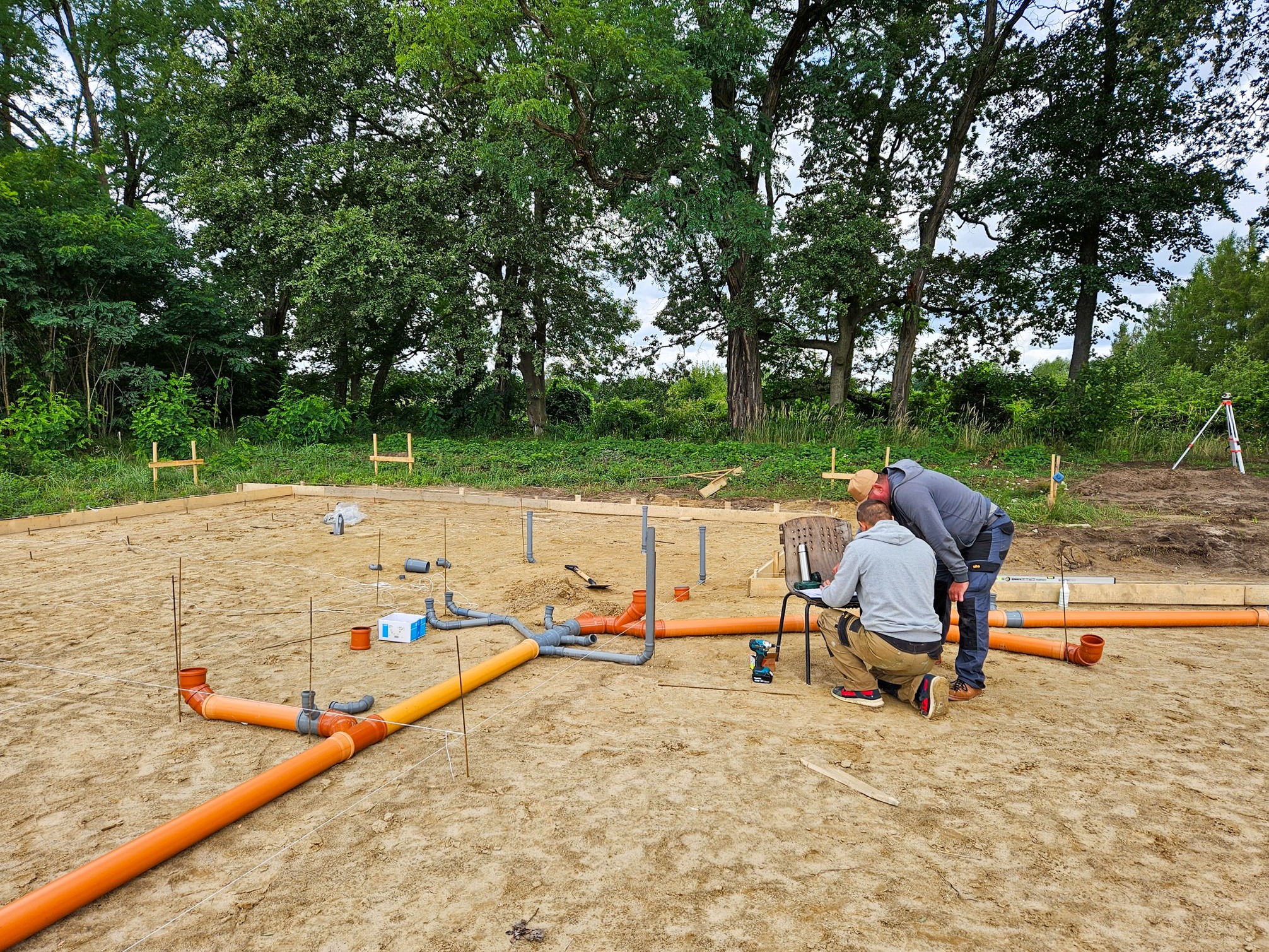 Dwaj pracownicy sprawdzają plan instalacji kanalizacyjnej na fundamencie domu szkieletowego, widoczne rury kanalizacyjne pomarańczowe i szare, poziomica oraz niwelator laserowy w tle.