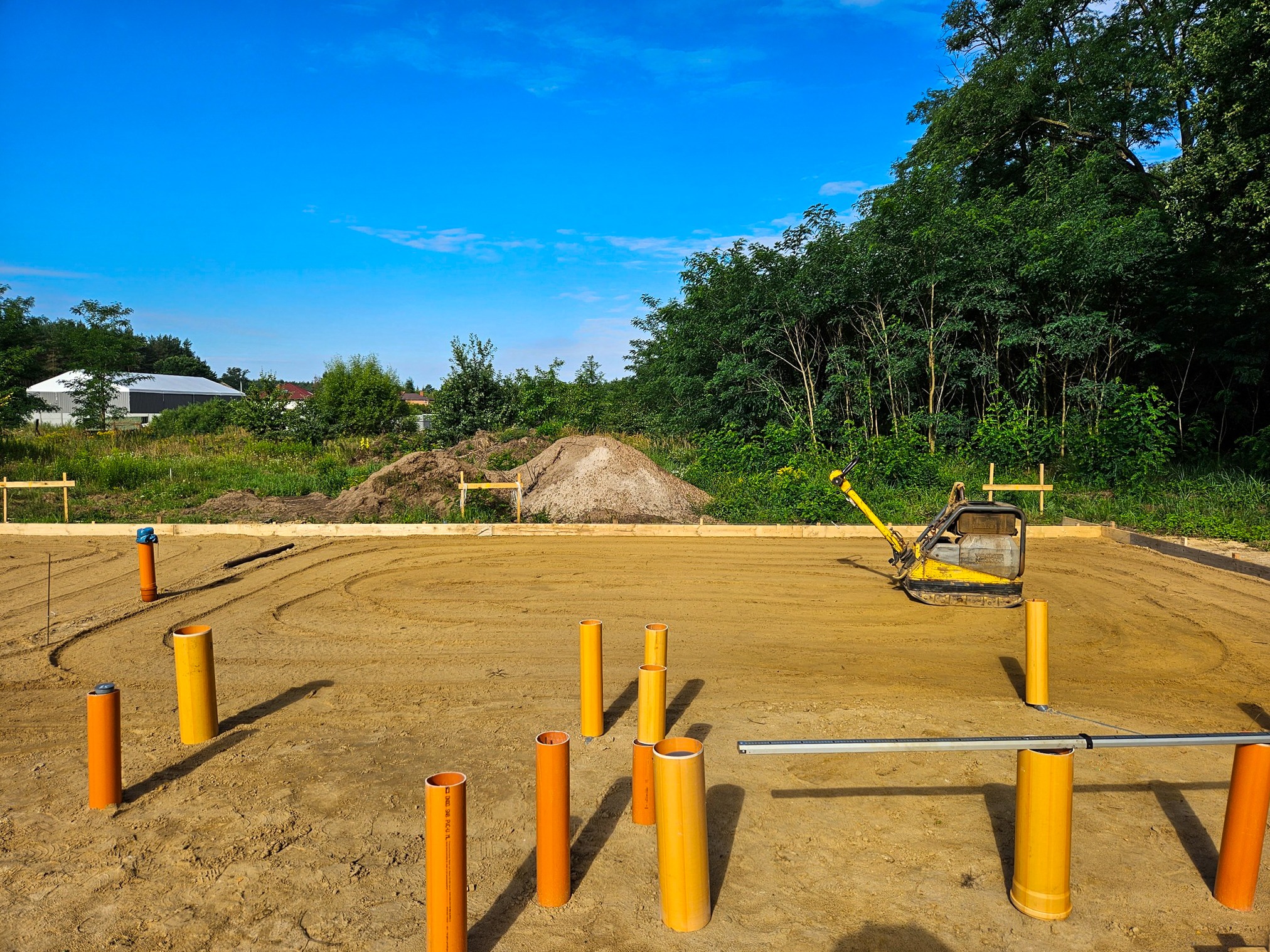 Przygotowanie terenu pod fundamenty: wyrównany teren z rurami kanalizacyjnymi i małą koparką, widok na budowę w słoneczny dzień.