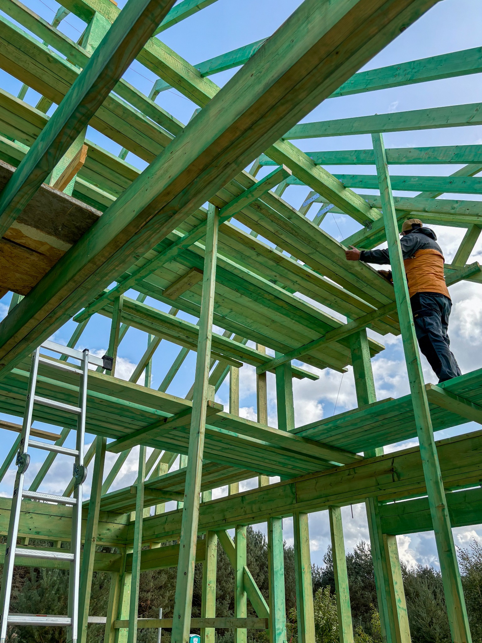 Konstrukcja szkieletowa domu w trakcie montażu, widoczny pracownik na wysokości mocujący elementy dachu, drabina oparta o konstrukcję, błękitne niebo w tle.