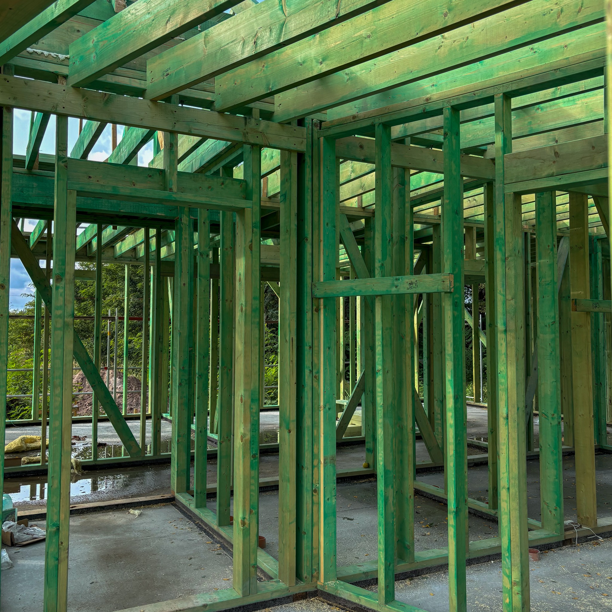 Wnętrze budynku w trakcie budowy, widoczna zielona drewniana konstrukcja szkieletowa z widocznymi belkami stropowymi i słupami, betonowa podłoga z kałużami wody.