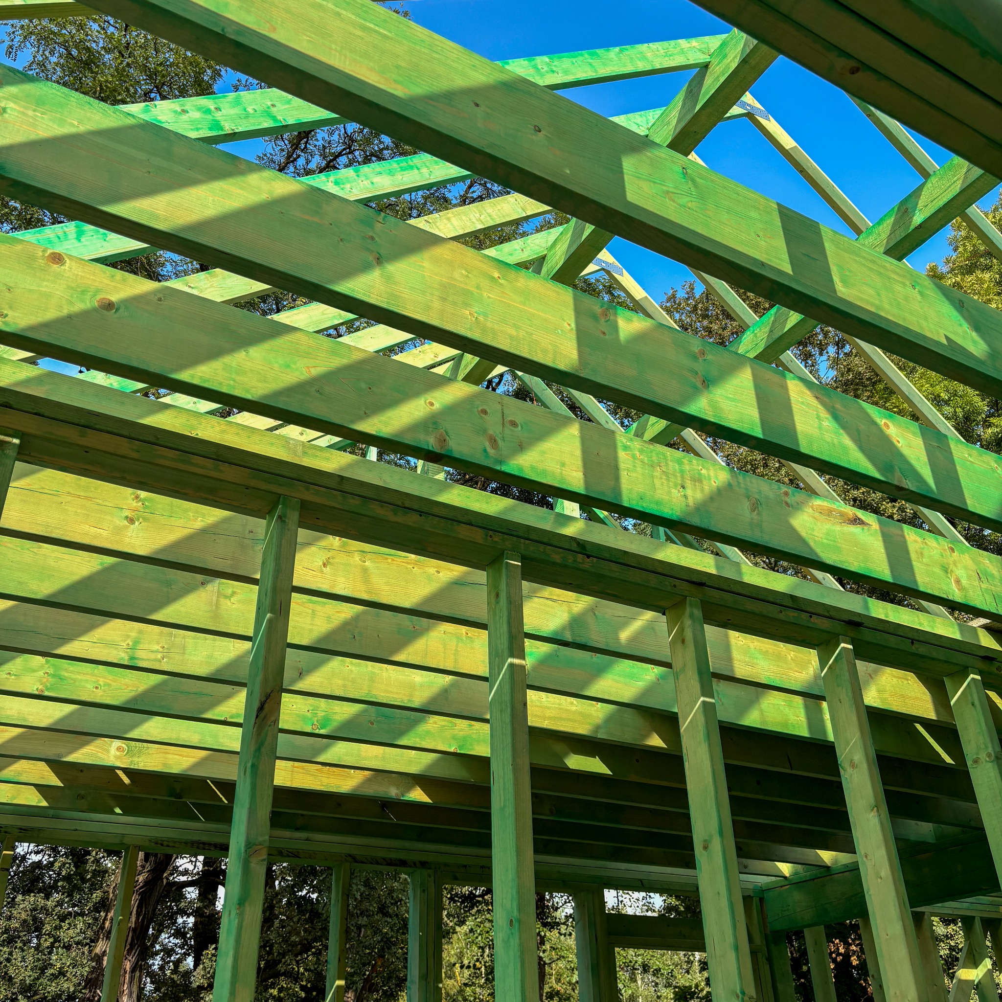 Wnętrze konstrukcji domu w trakcie budowy z belek pomalowanych na zielono, widok na niebo i drzewa przez otwory w konstrukcji.