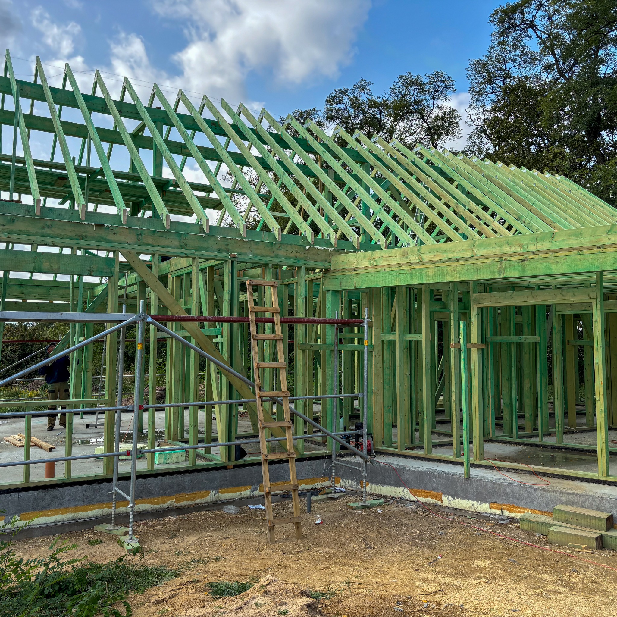 Konstrukcja szkieletowa domu w trakcie budowy, widoczne zielone drewniane elementy, rusztowanie i drabina, fundament z izolacją, częściowo pochmurne niebo w tle.