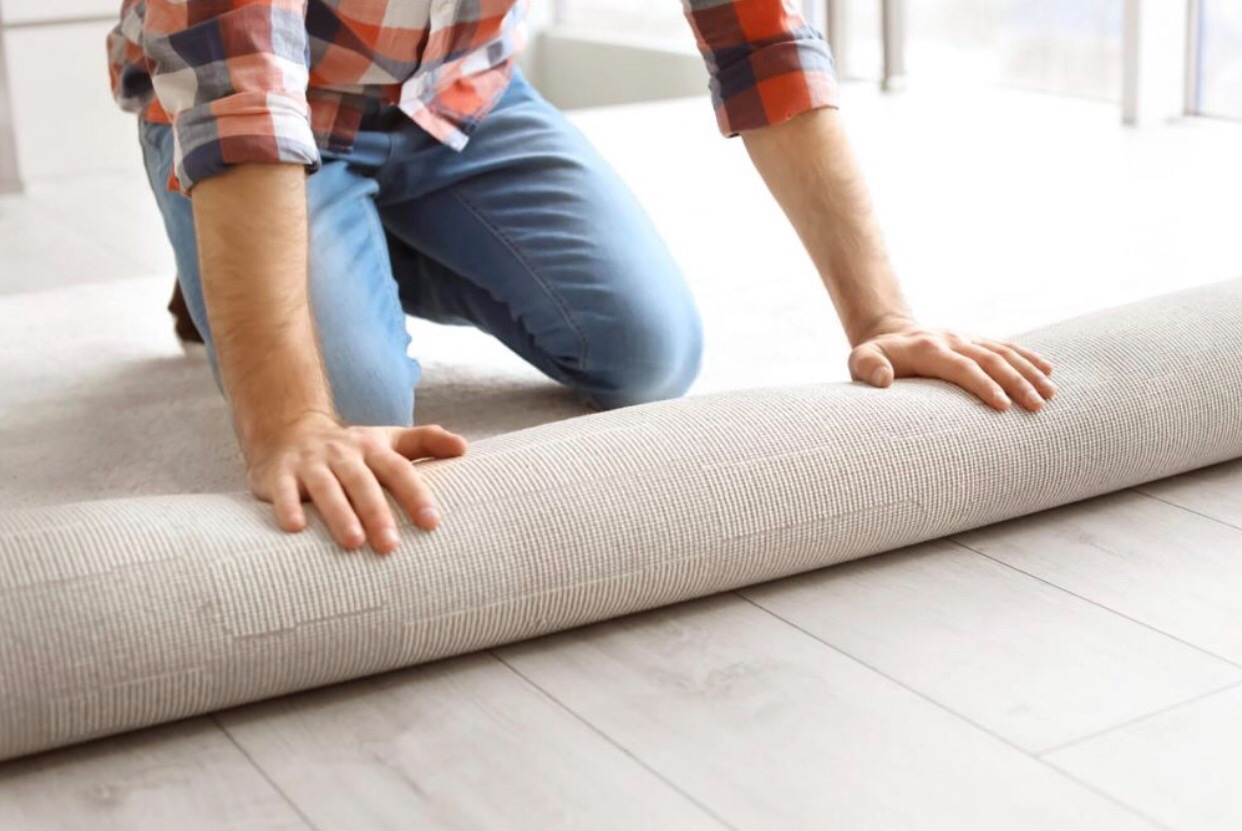 Mężczyzna w koszuli w kratę rozkłada zwiniętą, beżową wykładzinę na jasnej podłodze z paneli.