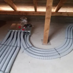 Szare, elastyczne rury wentylacyjne połączone z metalową skrzynką rozdzielczą, zamontowane w surowym wnętrzu poddasza z drewnianą konstrukcją dachu i betonową podłogą.