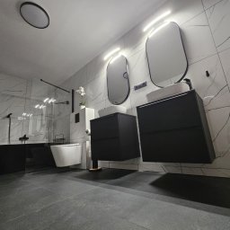 DrabMar - Nowoczesna łazienka z czarną wanną wolnostojącą, wiszącymi szafkami z umywalkami i białymi płytkami imitującymi marmur.