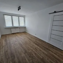 Pokój po remoncie z brązowymi panelami podłogowymi, białymi ścianami, grzejnikiem pod oknem z żaluzjami poziomymi i przesuwnymi drzwiami w stylu stodoły z czarnymi akcentami.