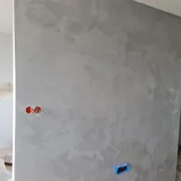 Ściana pokryta szarą, dekoracyjną powłoką z efektem chmur, widoczne puszki z farbą i narzędzia malarskie w tle, gniazdko elektryczne bez obudowy.